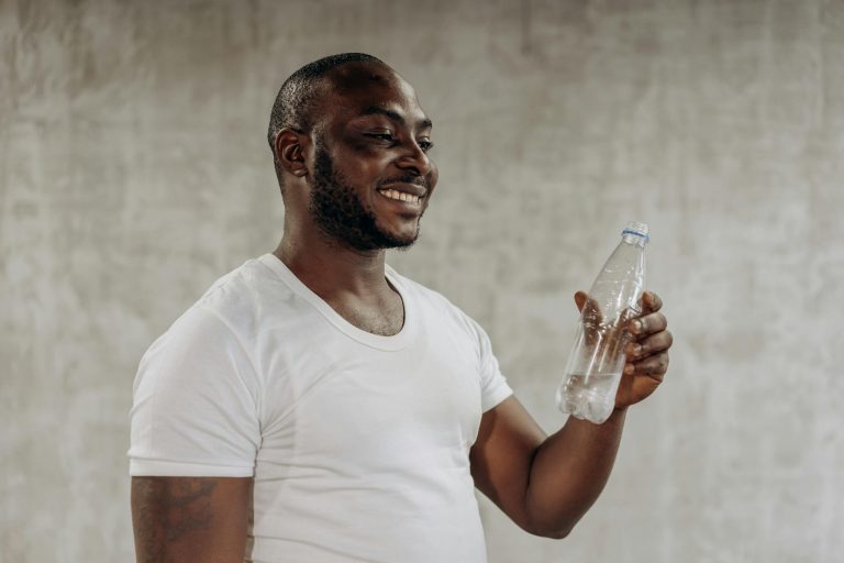 A happy man smiles while holding a bottle of water indoors, embracing healthy living.