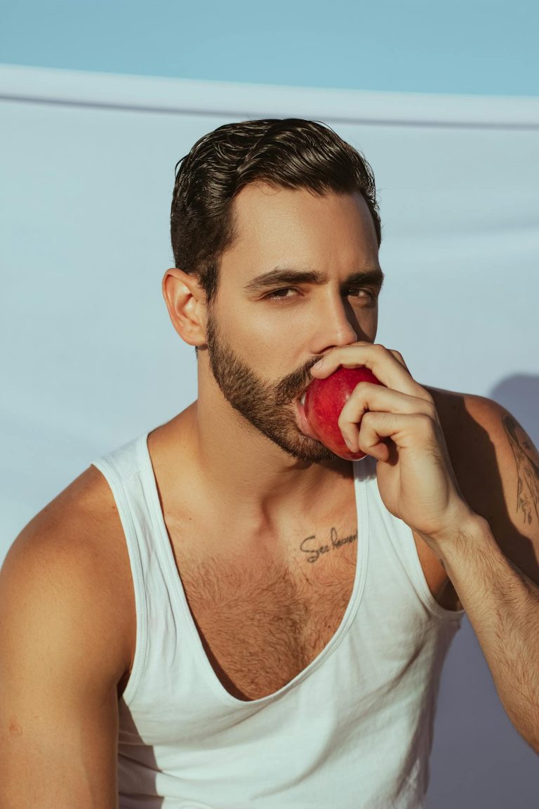 Stylish man in a white tank top enjoying a red apple under sunny skies.